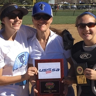 Three women on a softball field pose for a photo. The woman in the middle holds a USSSA plaque, and the woman on the right holds an MVP award. They are smiling and wearing athletic gear.