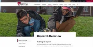 Two students work outdoors on a grassy slope at the University of Denver. One student examines the ground, while the other holds a laptop. The university's logo is visible at the top of the image, alongside navigation options for their website.