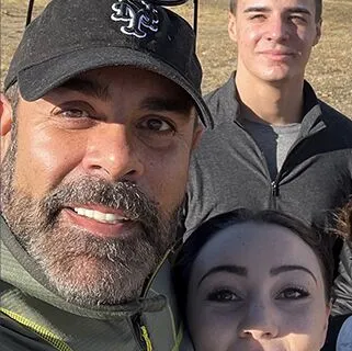 A group selfie with four people outdoors. The person in front is wearing a dark cap and jacket. They all appear to be smiling or content. A field is visible in the background under natural light.