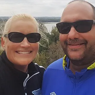 A woman and a man wearing sunglasses smile for a selfie outdoors. They are dressed in casual jackets and standing in front of a scenic view with trees and a body of water in the background.