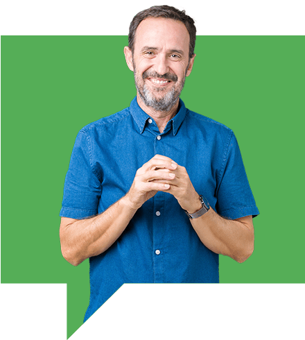 A man with a beard smiles while standing against a green background. He is wearing a short-sleeved blue button-up shirt and holding his hands together in front.