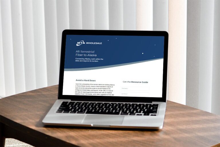 A laptop sits on a wooden table, displaying a webpage with a blue and white design. The text reads, "MTA Wholesale All-Terrestrial Fiber to Alaska." The page features sections titled "Avoid a Hard Down" and "Get the Resource Guide." Vertical blinds are in the background.