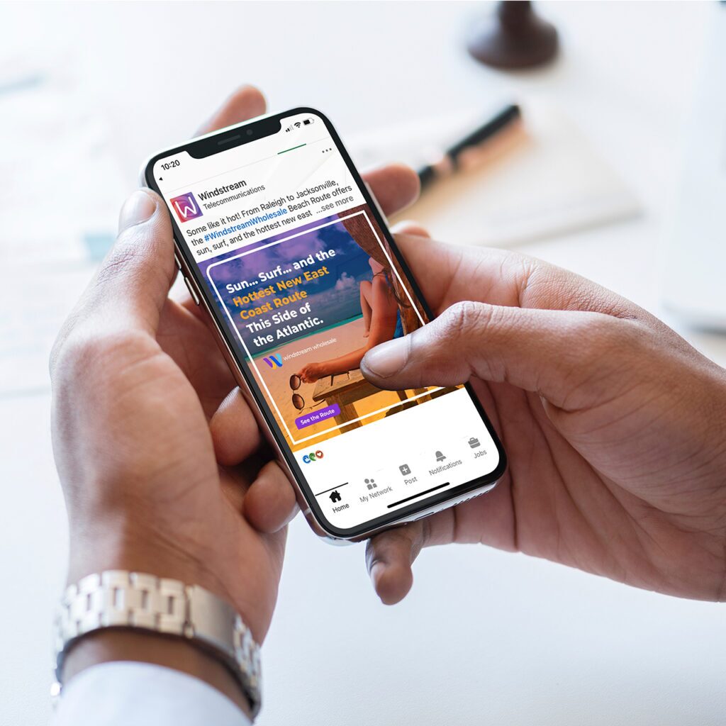A person holding a smartphone displaying a website or app. The screen shows an ad with text and an image of a beach scene, including sand and a surfboard. The background is blurred, with a pen and other items on a table.