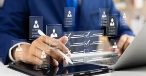 A person in a suit uses a stylus to interact with a digital tablet, surrounded by floating icons of LinkedIn profiles and checklists. A laptop is open nearby, hinting at activities related to recruitment or social selling habits.