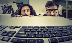 Two men with glasses and expressive eyes peer over a desk at a computer keyboard in the foreground. They appear curious and slightly anxious, with bookshelves and cushions in the background.