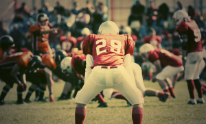 A football player in a red uniform, number 29, prepares for a play in a game. Opposing team players in orange are positioned in the background. Spectators sit in the stands, watching the action. The scene is set on a sunny day.