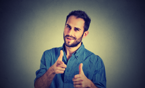 A man with a beard and short hair is smiling and pointing both index fingers forward. He wears a blue button-up shirt. The background is plain and slightly textured.