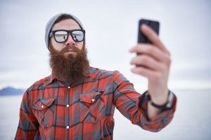 A bearded man wearing sunglasses, a gray beanie, and a red plaid shirt takes a selfie with his smartphone. He is standing outdoors with a snowy landscape in the background.