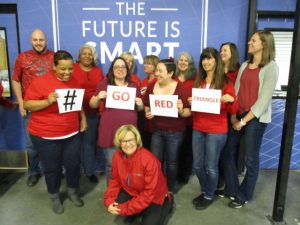A group of people wearing red stand together for a photo. Some are holding signs that read "#GO RED TRIANGLE." A banner in the background says "THE FUTURE IS SMART." A woman in a red jacket kneels in front.