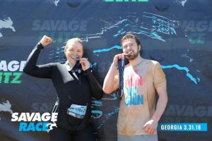 Two people celebrating at a Savage Race event. They are holding medals to their mouths. The woman is in black athletic wear, and the man in a muddy t-shirt. The background features the Savage Race logo. The text reads "Savage Blitz" and "Georgia 3.31.18".