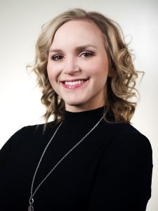 A person with blonde, curly hair smiles at the camera. They are wearing a black top and a silver necklace. The background is a plain, light color.