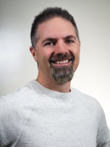 A man with short dark hair and a goatee smiles at the camera. He is wearing a light gray sweater against a plain background.