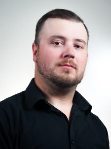 A man with short hair and a beard is wearing a black shirt. He is looking directly at the camera against a plain, light-colored background.