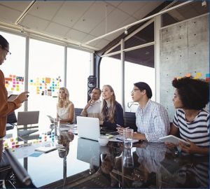 A diverse group of people are in a modern office, seated around a glass table with laptops and notepads. They're looking attentively at someone presenting. Colorful sticky notes cover a window in the background.