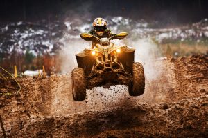 A person wearing a helmet and protective gear rides an ATV, airborne over a muddy track. The ATV's headlights are on, and dirt is flying around, creating an action-packed scene in a rugged outdoor setting.