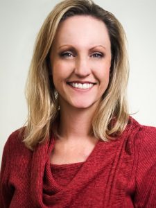 A person with shoulder-length blonde hair is smiling, wearing a red sweater against a neutral background.