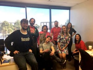 A group of nine people posing in an office with a large window. Some are seated and others standing, wearing casual and colorful attire. There are trees visible outside the window. Everyone is smiling.