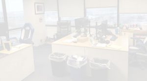 An office with two desks, each equipped with computers and chairs. There are large windows with blinds partially open, allowing natural light to enter. Two trash cans are placed between the desks, and various office supplies are scattered around.