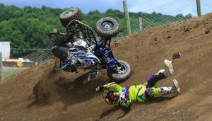 A person in bright green and purple motocross gear falls off their blue ATV, which is airborne, during a dirt track race. The rider is sliding on the ground, surrounded by dirt with a green forest in the background.