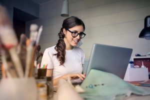 A woman wearing glasses and a white t-shirt is sitting at a desk, smiling while using a laptop. In the foreground, there are art supplies and papers, suggesting a creative workspace. The room is softly lit.