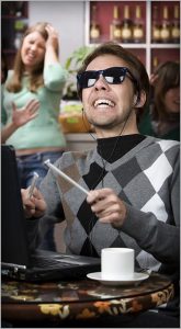 A person wearing dark sunglasses and headphones is enthusiastically playing a laptop keyboard like drums, with a joyful expression. A white cup is on the table. In the background, a group of people is talking and smiling.