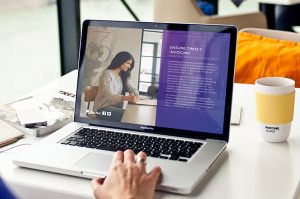 A person using a MacBook Pro displaying a webpage about ensuring timely invoicing. The screen shows an image of a woman writing and text on a purple background. In the foreground, a hand rests on the laptop and a Pantone mug is nearby.
