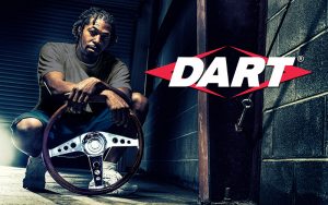 A man crouches in a dimly lit garage, holding a steering wheel. The word "DART" is prominently displayed in bold letters. The setting has industrial elements, with a brick wall in the background.