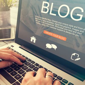 A person is typing on a laptop with a screen displaying a blog creation page. Icons for home, speech bubbles, and a pencil are visible, along with the "Create Your Blog" button. The scene is well-lit, emphasizing the keyboard and display.
