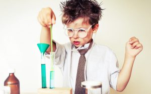 A young boy with messy hair and glasses, wearing a white shirt and tie, uses a pipette with green liquid in a test tube. He stands in a playful setup resembling a scientist's lab, experimenting as though he's cracking the code to Marketing Automation with bottles surrounding him.