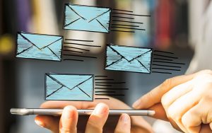 A person's hand uses a smartphone, with illustrated blue envelopes symbolizing emails floating above it, indicating digital communication or email exchange.