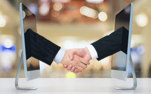 Two people shaking hands through computer screens, symbolizing a virtual agreement or online partnership. The background is blurred, highlighting the focus on the handshake and digital communication.