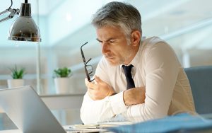 A man with gray hair in a shirt and tie holds glasses, staring thoughtfully at a laptop on a desk. There are papers, a lamp, and plants visible. The setting appears to be a modern office with large windows.