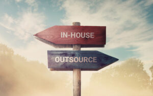 A wooden signpost in a foggy landscape displays two arrows: one red, labeled "IN-HOUSE," pointing left, and one blue, labeled "OUTSOURCE," pointing right. The background features a cloudy sky and silhouettes of trees.