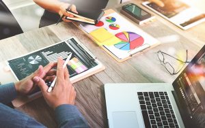 Two people collaborate at a desk with charts and graphs on clipboards, a laptop, and a smartphone. One, possibly a marketing consultant, gestures with a pen while the other takes notes. The scene conveys an analysis session or business meeting.