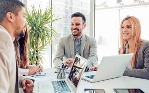 Four people are sitting around a table in a bright office with large windows, discussing sales enablement strategies. They are smiling and using laptops, while one person diligently takes notes. A green plant adds a touch of nature to the vibrant setting.