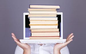 A person emerging from a laptop is holding a stack of books, symbolizing digital education and webinar best practices. The individual, sporting a white shirt, has only their hands and part of the shirt visible.