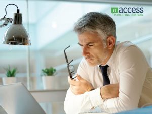 A man with gray hair sits at a desk, intently looking at a computer screen while holding his glasses. He’s dressed in a white shirt and tie. The background features a modern office space, with the logo "Access Marketing Company" visible in the corner.