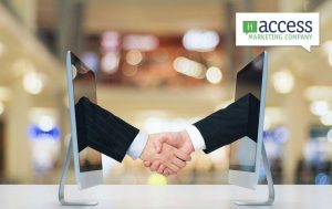 Two computer monitors face each other on a table, with a handshake appearing between them. A logo in the top right corner reads "Access Marketing Company." The background is a blurred interior space with warm lighting.