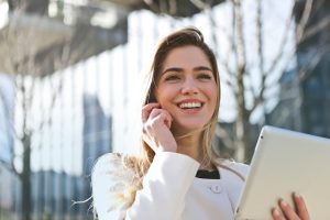 A smiling woman with long hair is outdoors, holding a tablet and talking on her phone. She is wearing a light-colored coat, and the background features blurred buildings and trees on a sunny day.