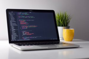 A laptop on a white desk displays code on its screen. Next to it is a yellow mug with a floral pattern. In the background, there is a green potted plant.