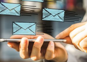 A person holds a smartphone with several cartoon-style blue envelopes overlaid, suggesting emails or messages being sent. The background is blurred.
