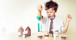 A young child in glasses and a tie is conducting an experiment with test tubes and colorful liquids, reminiscent of the intricate processes behind marketing automation. Various bottles and jars are on the table as the child appears focused and curious.