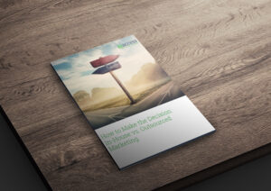 A brochure rests on a wooden table, featuring a cover image of a crossroads with a blank signpost under a cloudy sky. The title reads, "How to Make the Decision: In-House vs. Outsourced Marketing.