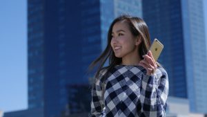 A woman wearing a plaid sweater holds a smartphone and smiles against a backdrop of tall modern buildings and a clear blue sky.