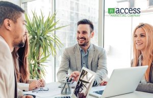 A group of four people in business attire sits around a table with laptops and documents, engaged in a discussion about sales enablement. They smile at each other, while a large plant and a window with a city view create an inspiring backdrop. The logo reads "Access Marketing Company.