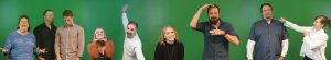A group of seven people standing against a green background, each striking different playful or expressive poses. They are wearing casual to semi-formal attire and are positioned in a horizontal line.