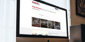 Close-up of a computer screen displaying a website with a search bar and images related to communication and conferencing. The screen is set in a dimly lit room with a window on the left and a framed picture on the right.