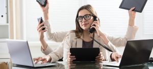 A woman with multiple arms is multitasking at a desk. She is holding a phone, tablet, calculator, and smartphone while using a laptop. She sits in an office setting with blinds on the windows behind her.