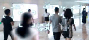 A blurred image of six people walking through a brightly lit corridor with digital overlays indicating estimated age and gender: "Male Age 21," "Female Age 23," "Male Age 29," and "Female Age 35.