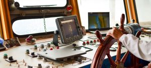 A person at the helm of a ship navigates using various control instruments. The scene includes a wooden steering wheel, multiple control buttons, and various screens displaying navigational data, set against a window overlooking the ocean.